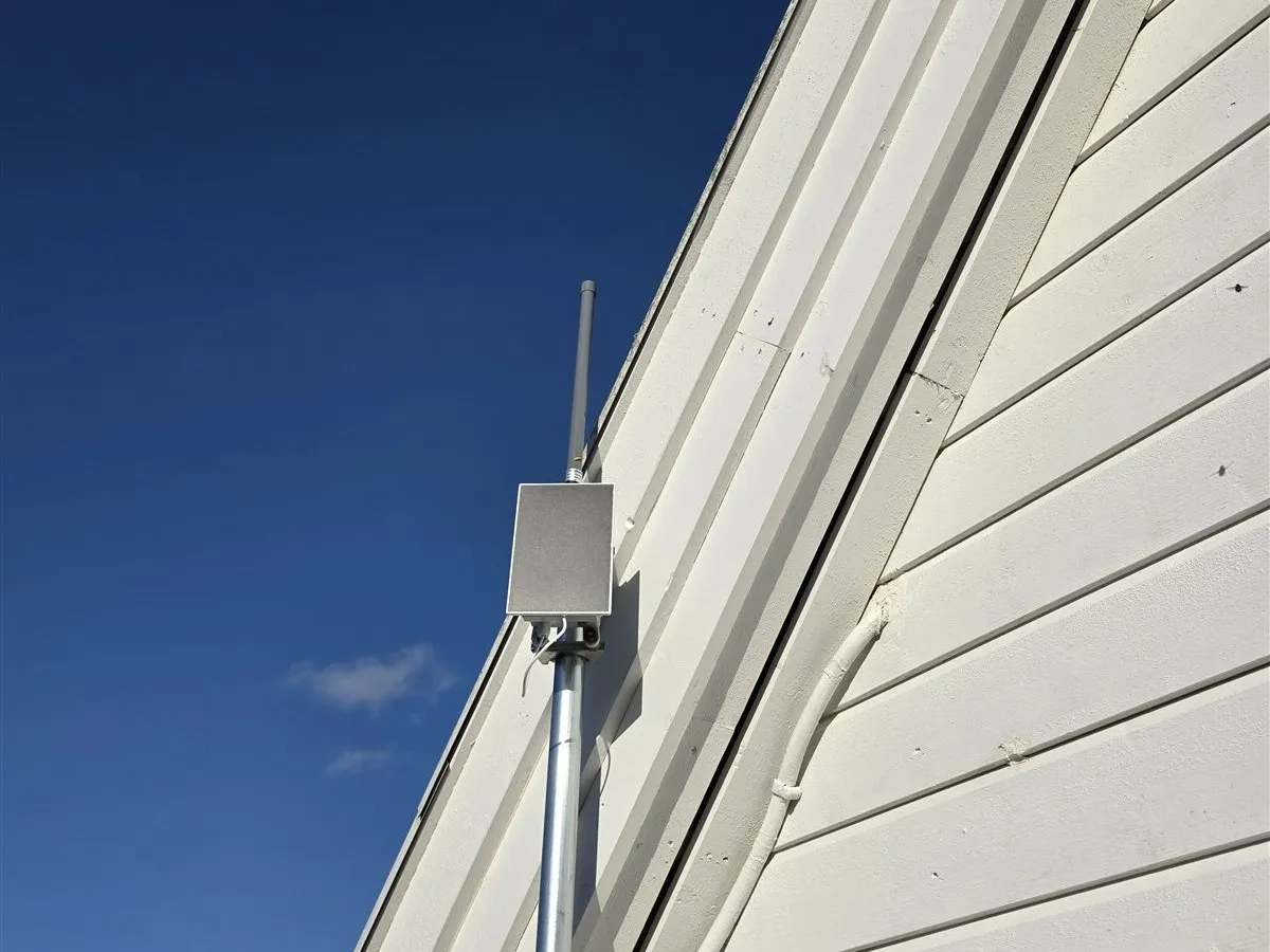 The MeshCore repeater in a small grey box with an antenna, on a pole by the gable of the house